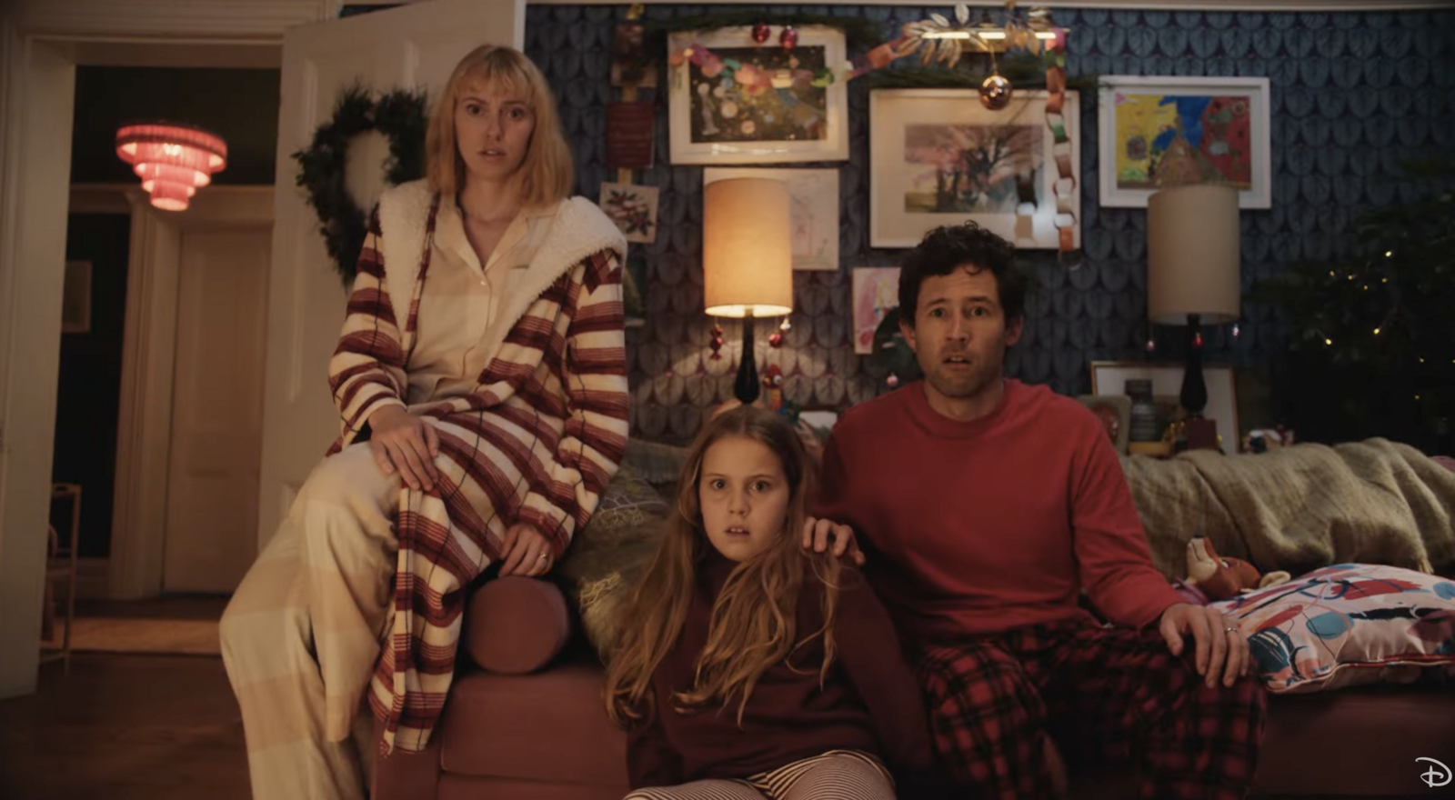 Une femme, un homme et une jeune fille sont assis sur le canapé d'un salon, l'air surpris. La pièce est décorée d'œuvres d'art et de couronnes de Noël pour Le Plus Beau Des Noëls, inspiré de la magie du Court-métrage Noël Disney.