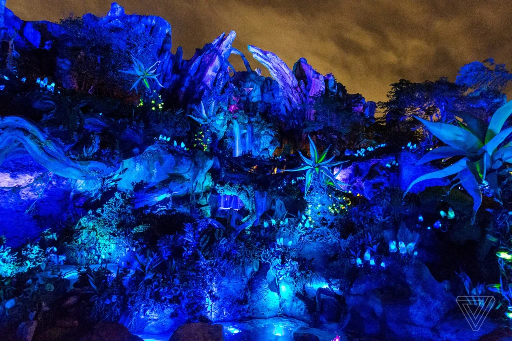 Scène nocturne dans Avatar Land, où des lumières bleues et violettes illuminent des rochers et des plantes d'apparence extraterrestre sous un ciel nuageux - une nouvelle attraction de Disneyland Resort.