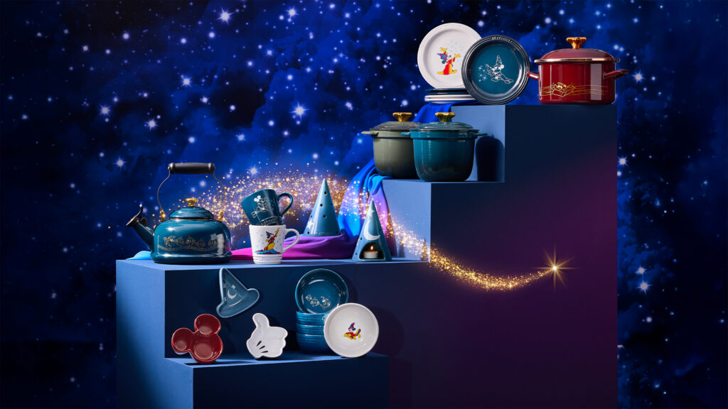 Articles de cuisine assortis sur le thème de Disney, notamment des théières Fantasia, des ustensiles de cuisine Le Creuset, des assiettes, des tasses et des ustensiles Disney, présentés sur des marches bleues sur un fond étoilé avec des paillettes.