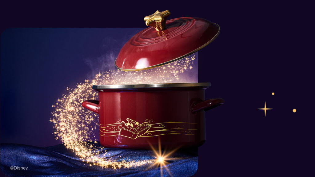 Un pot Le Creuset à couvercle rouge avec des poignées dorées et un motif décoratif libère de la vapeur et scintille sur un tissu bleu, évoquant la magie de Fantasia sur un fond sombre.