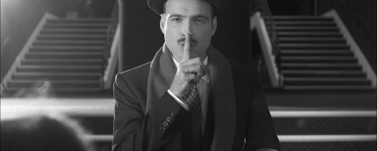 Un homme en costume et chapeau se tient devant les escaliers du Théâtre du Lucernaire à Paris, portant un doigt à ses lèvres en signe de "chut".