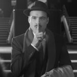 Un homme en costume et chapeau se tient devant les escaliers du Théâtre du Lucernaire à Paris, portant un doigt à ses lèvres en signe de "chut".