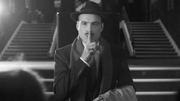 Un homme en costume et chapeau se tient devant les escaliers du Théâtre du Lucernaire à Paris, portant un doigt à ses lèvres en signe de "chut".