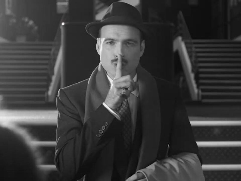 Un homme en costume et chapeau se tient devant les escaliers du Théâtre du Lucernaire à Paris, portant un doigt à ses lèvres en signe de "chut".