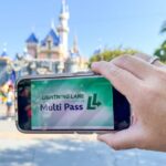 Une main tient un téléphone affichant l'écran "Lightning Lane Multi Pass" devant l'emblématique château de Disneyland, capturant l'excitation de l'entrée anticipée dans le parc à thème.
