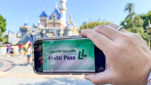 Une main tient un téléphone affichant l'écran "Lightning Lane Multi Pass" devant l'emblématique château de Disneyland, capturant l'excitation de l'entrée anticipée dans le parc à thème.