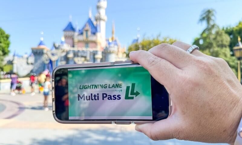 Une main tient un téléphone affichant l'écran "Lightning Lane Multi Pass" devant l'emblématique château de Disneyland, capturant l'excitation de l'entrée anticipée dans le parc à thème.