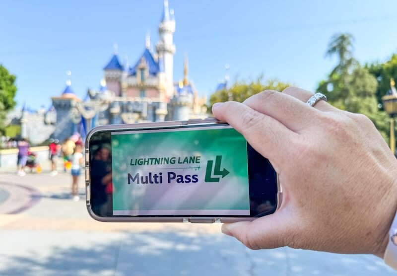 Une main tient un téléphone affichant l'écran "Lightning Lane Multi Pass" devant l'emblématique château de Disneyland, capturant l'excitation de l'entrée anticipée dans le parc à thème.