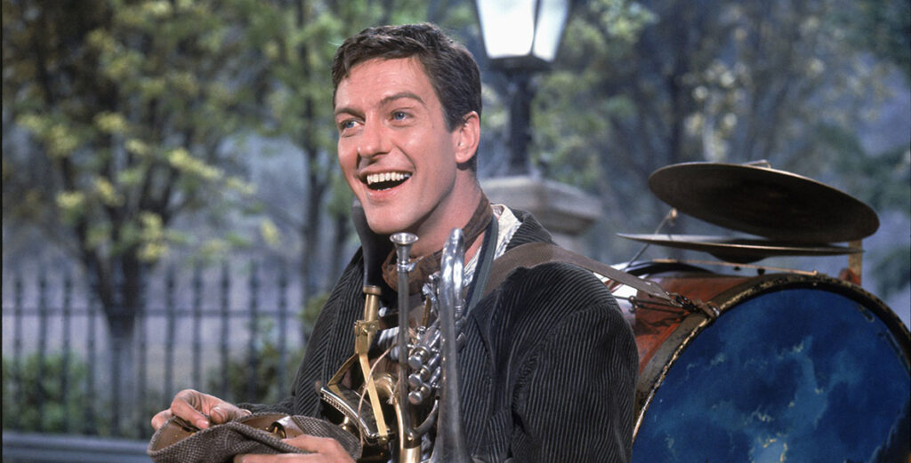 Un homme en plein air sourit tout en portant une tenue d'homme-orchestre rappelant le personnage de Dick Van Dyke dans Mary Poppins. Divers instruments de musique sont attachés à son corps, avec des arbres et un lampadaire en arrière-plan.
