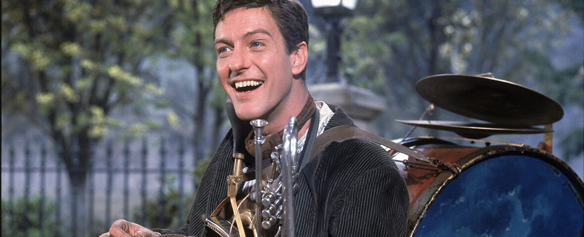 Un homme en plein air sourit tout en portant une tenue d'homme-orchestre rappelant le personnage de Dick Van Dyke dans Mary Poppins. Divers instruments de musique sont attachés à son corps, avec des arbres et un lampadaire en arrière-plan.