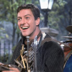 Un homme en plein air sourit tout en portant une tenue d'homme-orchestre rappelant le personnage de Dick Van Dyke dans Mary Poppins. Divers instruments de musique sont attachés à son corps, avec des arbres et un lampadaire en arrière-plan.
