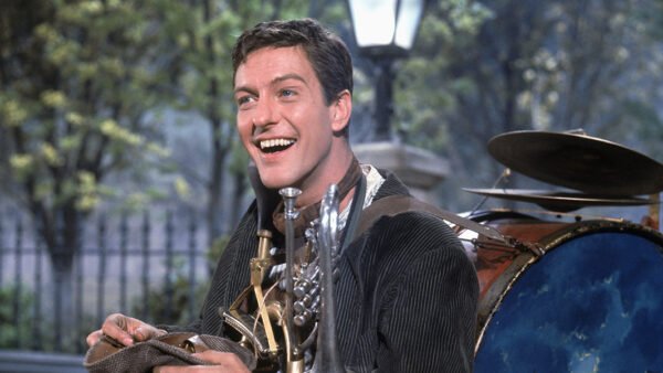 Un homme en plein air sourit tout en portant une tenue d'homme-orchestre rappelant le personnage de Dick Van Dyke dans Mary Poppins. Divers instruments de musique sont attachés à son corps, avec des arbres et un lampadaire en arrière-plan.