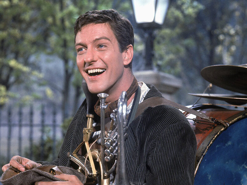 Un homme en plein air sourit tout en portant une tenue d'homme-orchestre rappelant le personnage de Dick Van Dyke dans Mary Poppins. Divers instruments de musique sont attachés à son corps, avec des arbres et un lampadaire en arrière-plan.