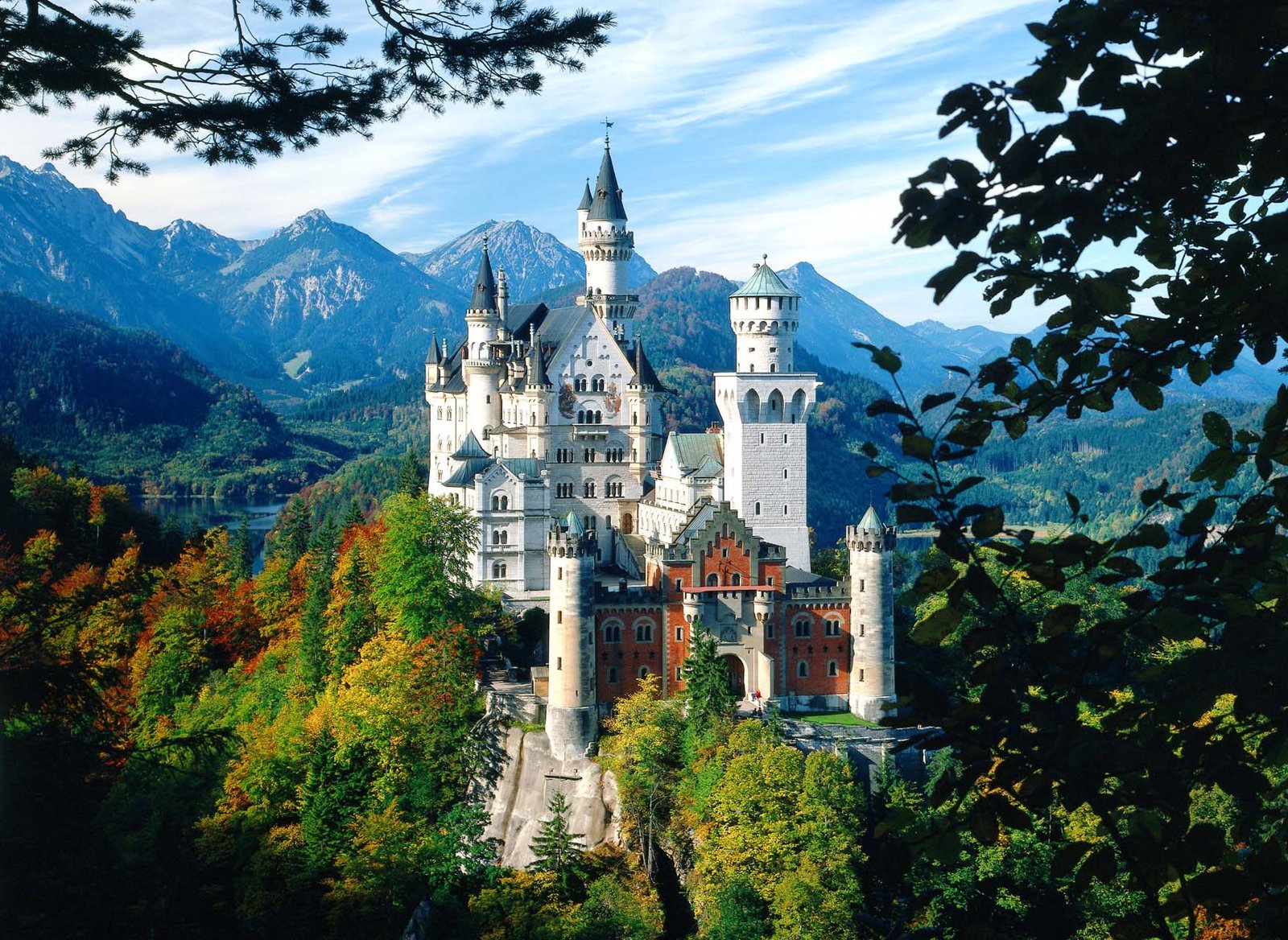 Le château de Neuschwanstein, qui rappelle un château de conte de fées et qui aurait inspiré La Belle au bois dormant de Disney, se dresse au sommet d'une colline boisée avec des montagnes en arrière-plan et des arbres au premier plan par temps clair.