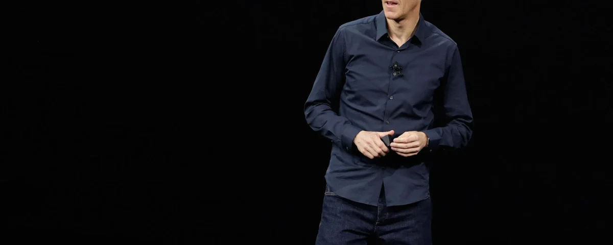 Un homme vêtu d'une chemise sombre et d'un jean se tient sur scène sur un fond noir, canalisant sa technologie d'expertise tout en parlant et en faisant des gestes avec ses mains.