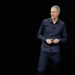 Un homme vêtu d'une chemise sombre et d'un jean se tient sur scène sur un fond noir, canalisant sa technologie d'expertise tout en parlant et en faisant des gestes avec ses mains.