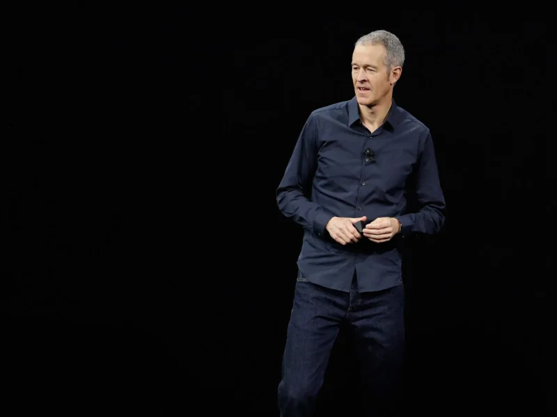Un homme vêtu d'une chemise sombre et d'un jean se tient sur scène sur un fond noir, canalisant sa technologie d'expertise tout en parlant et en faisant des gestes avec ses mains.