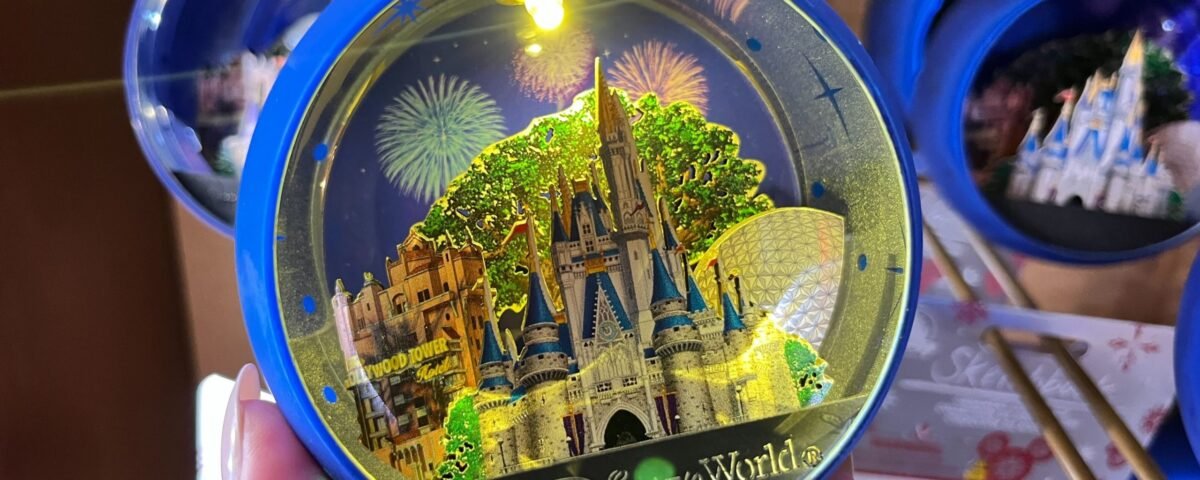 Une main tient un ornement bleu de Walt Disney World représentant le château de Cendrillon, Spaceship Earth et des feux d'artifice - l'un des souvenirs incontournables du parc - tandis que des ornements et des emballages similaires sont visibles à l'arrière-plan.