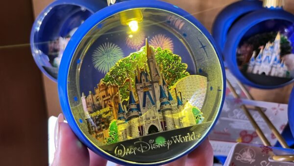Une main tient un ornement bleu de Walt Disney World représentant le château de Cendrillon, Spaceship Earth et des feux d'artifice - l'un des souvenirs incontournables du parc - tandis que des ornements et des emballages similaires sont visibles à l'arrière-plan.