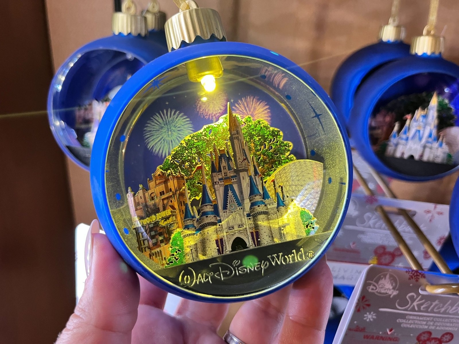 Une main tient un ornement bleu de Walt Disney World représentant le château de Cendrillon, Spaceship Earth et des feux d'artifice - l'un des souvenirs incontournables du parc - tandis que des ornements et des emballages similaires sont visibles à l'arrière-plan.
