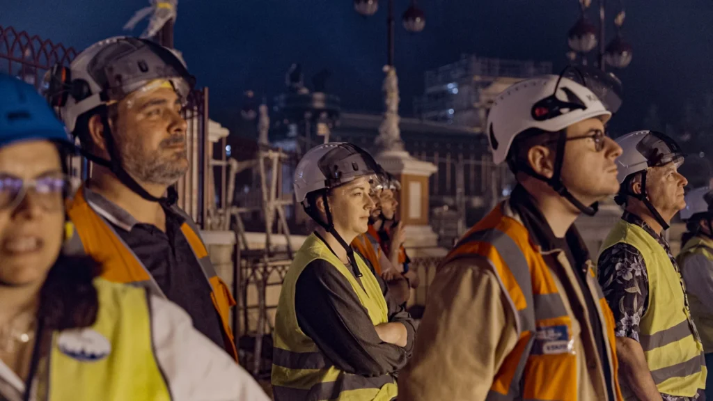 Un groupe d'ouvriers du bâtiment portant des casques et des gilets de sécurité se tient à l'extérieur, la nuit, et contemple avec émerveillement la cascade de lumières de Disney, captivé par l'éblouissant Spectacle Nocturne qui se déroule devant eux.