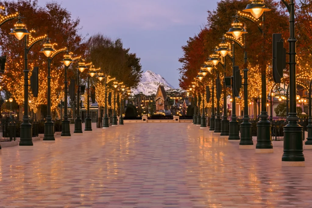Une large allée vide bordée de lampadaires illuminés et d'arbres couverts de guirlandes lumineuses mène vers une montagne enneigée au crépuscule, évoquant la magie de Disney Adventure World sur une musique originale de Philippe Rombi.