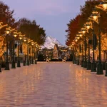 Une large allée vide bordée de lampadaires illuminés et d'arbres couverts de guirlandes lumineuses mène vers une montagne enneigée au crépuscule, évoquant la magie de Disney Adventure World sur une musique originale de Philippe Rombi.