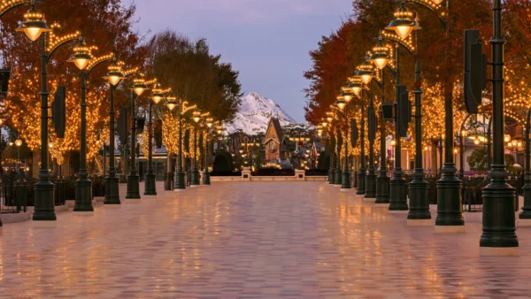Une large allée vide bordée de lampadaires illuminés et d'arbres couverts de guirlandes lumineuses mène vers une montagne enneigée au crépuscule, évoquant la magie de Disney Adventure World sur une musique originale de Philippe Rombi.