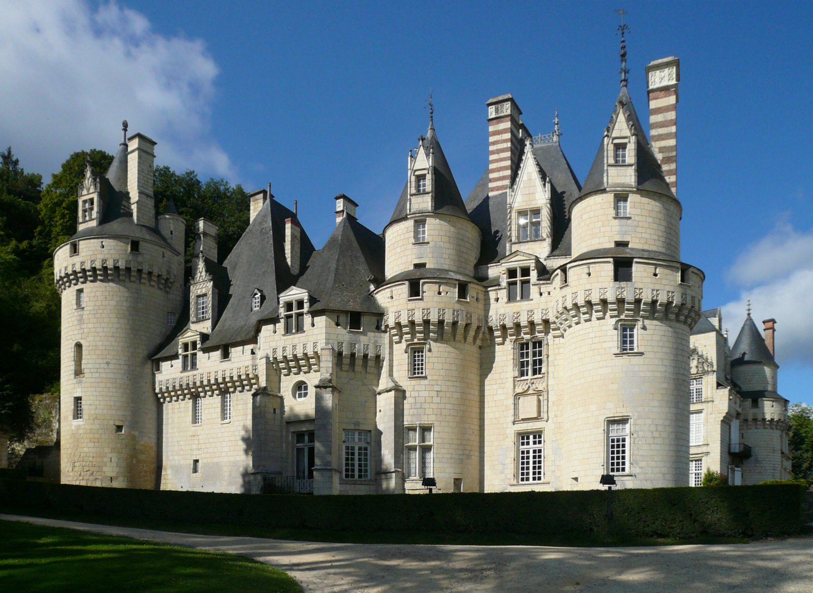 Un grand château en pierre avec de multiples tours rondes, des toits coniques et des cheminées décoratives évoque La Belle au Bois Dormant de Disney, sur fond de ciel bleu avec de la verdure à proximité.