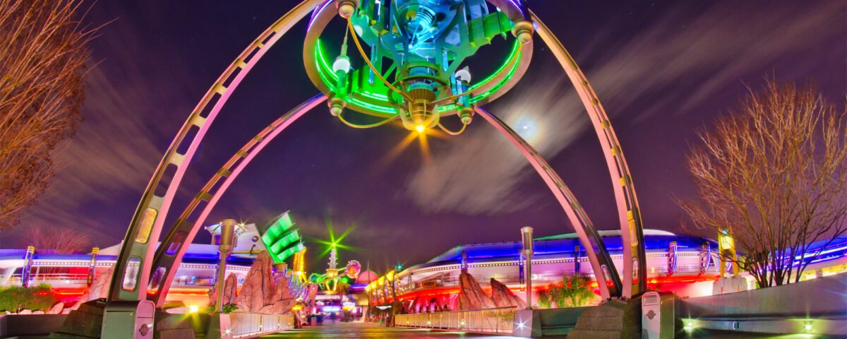L'entrée de Tomorrowland, de nuit, avec une grande enseigne lumineuse et des arches futuristes aux lumières colorées, illustre l'esprit d'innovation de Disney, avec un monorail glissant en arrière-plan.