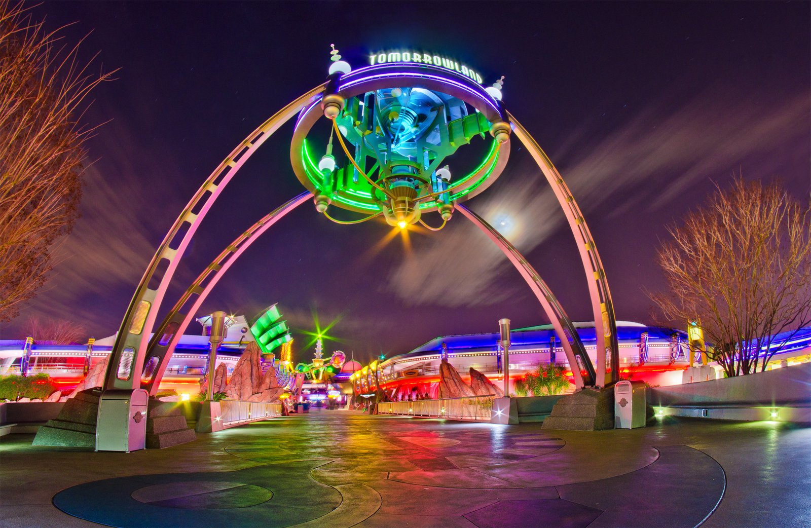 L'entrée de Tomorrowland, de nuit, avec une grande enseigne lumineuse et des arches futuristes aux lumières colorées, illustre l'esprit d'innovation de Disney, avec un monorail glissant en arrière-plan.