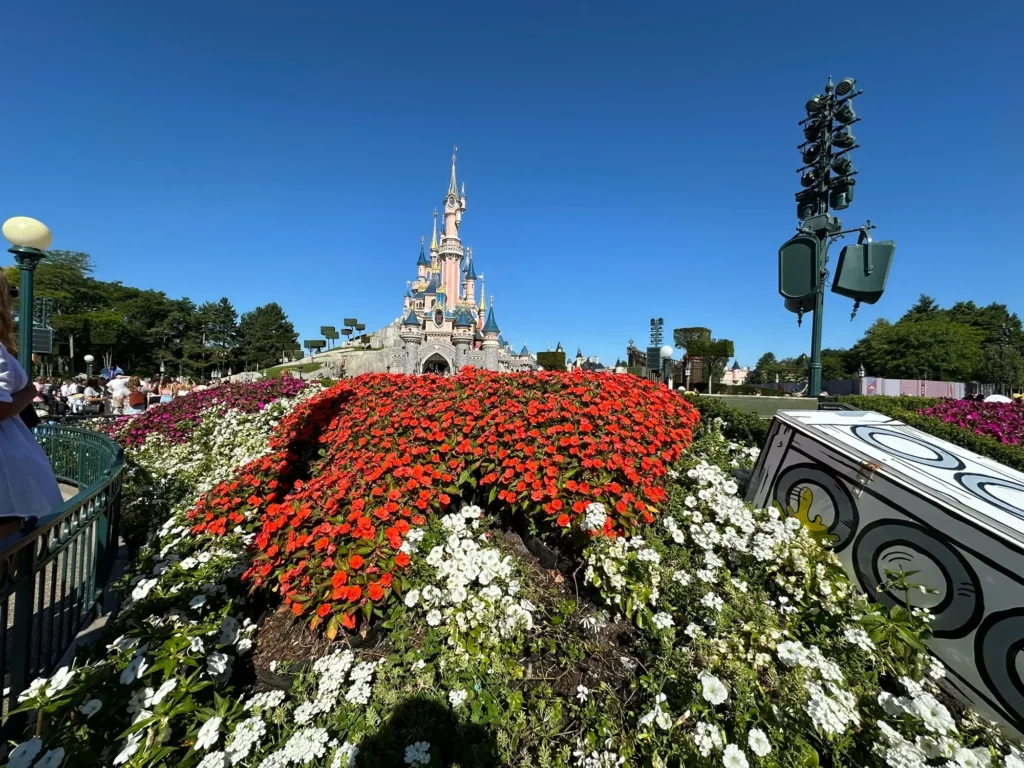 Un parterre de fleurs rouges et blanches devant un château de style féerique sous un ciel bleu clair à Disneyland Paris, où l'on peut s'initier à la biodiversité dans un cadre magique.