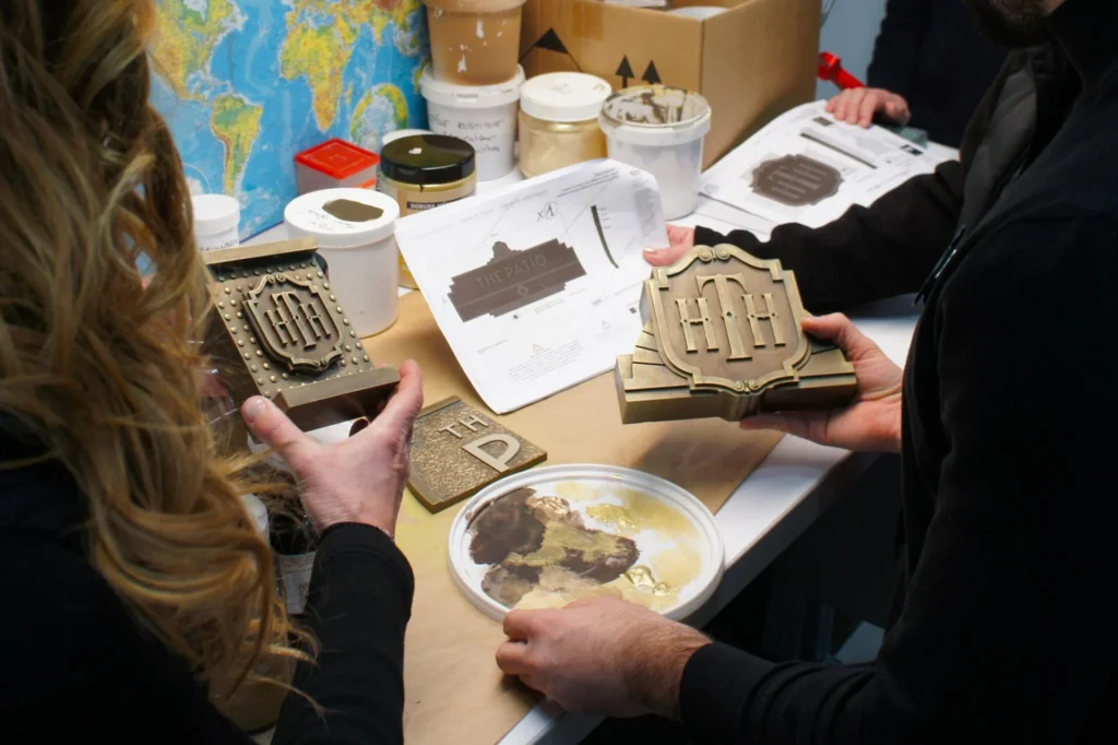 Deux personnes examinent des plaques métalliques et des échantillons de peinture sur une table avec, en arrière-plan, des documents de conception, des récipients de peinture et une carte du monde - peut-être des détails de planification pour Disneyland Paris ou le nouveau Disney Adventure World.