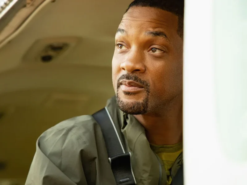 Un homme vêtu d'une veste verte avec une sangle sur l'épaule regarde pensivement par la portière ouverte d'un véhicule, une douce lumière naturelle illuminant son visage, évoquant l'esprit d'aventure de Nat Geo sur Disney+.