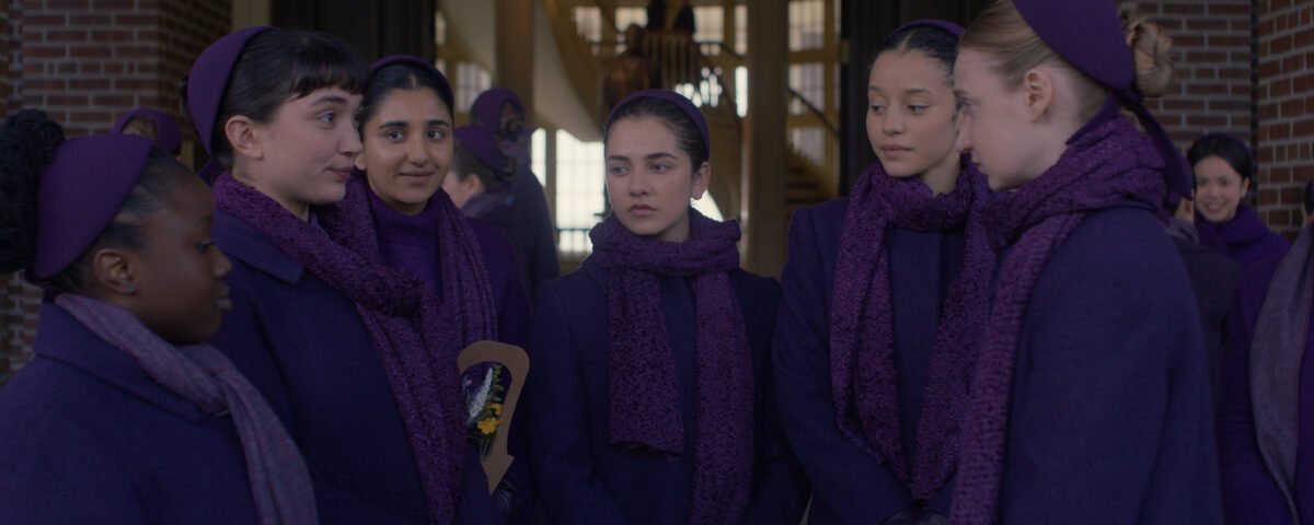 Un groupe de filles vêtues de manteaux, d'écharpes et de bandeaux violets assortis se tiennent ensemble et discutent dans un couloir, rappelant des scènes du spin-off de The Handmaid's Tale, The Testaments, qui sera bientôt disponible sur Disney+. D'autres personnes se mêlent à l'arrière-plan.