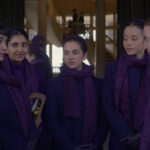 Un groupe de filles vêtues de manteaux, d'écharpes et de bandeaux violets assortis se tiennent ensemble et discutent dans un couloir, rappelant des scènes du spin-off de The Handmaid's Tale, The Testaments, qui sera bientôt disponible sur Disney+. D'autres personnes se mêlent à l'arrière-plan.