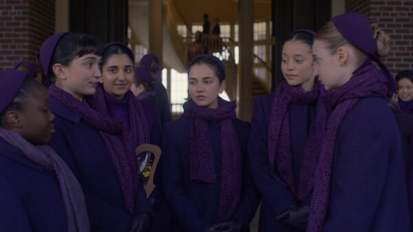 Un groupe de filles vêtues de manteaux, d'écharpes et de bandeaux violets assortis se tiennent ensemble et discutent dans un couloir, rappelant des scènes du spin-off de The Handmaid's Tale, The Testaments, qui sera bientôt disponible sur Disney+. D'autres personnes se mêlent à l'arrière-plan.