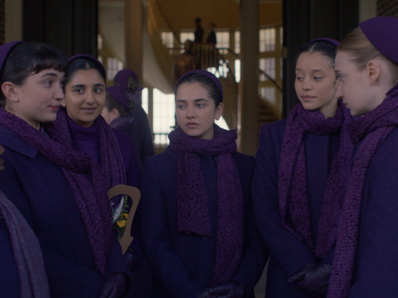 Un groupe de filles vêtues de manteaux, d'écharpes et de bandeaux violets assortis se tiennent ensemble et discutent dans un couloir, rappelant des scènes du spin-off de The Handmaid's Tale, The Testaments, qui sera bientôt disponible sur Disney+. D'autres personnes se mêlent à l'arrière-plan.