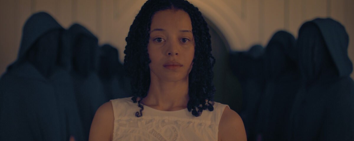 Une femme aux cheveux bouclés vêtue d'une robe blanche se tient au premier plan, entourée de personnes vêtues de robes sombres à capuchon, dans une pièce faiblement éclairée de The Testaments, le prochain spin-off de The Handmaid's Tale (La Servante) sur Disney+.