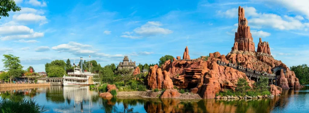 Une montagne rocheuse de style occidental avec une piste de montagnes russes se trouve à côté d'une rivière calme avec un bateau à aubes et des arbres sous un ciel bleu à Disneyland Paris.