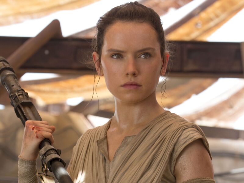 Une femme vêtue d'une tenue bronzée, rappelant le rôle emblématique de Daisy Ridley, se tient debout en tenant un bâton et regarde devant elle avec détermination dans un décor rustique et ensoleillé, évoquant l'esprit du Nouvel Ordre Jedi de La Guerre des étoiles 10.