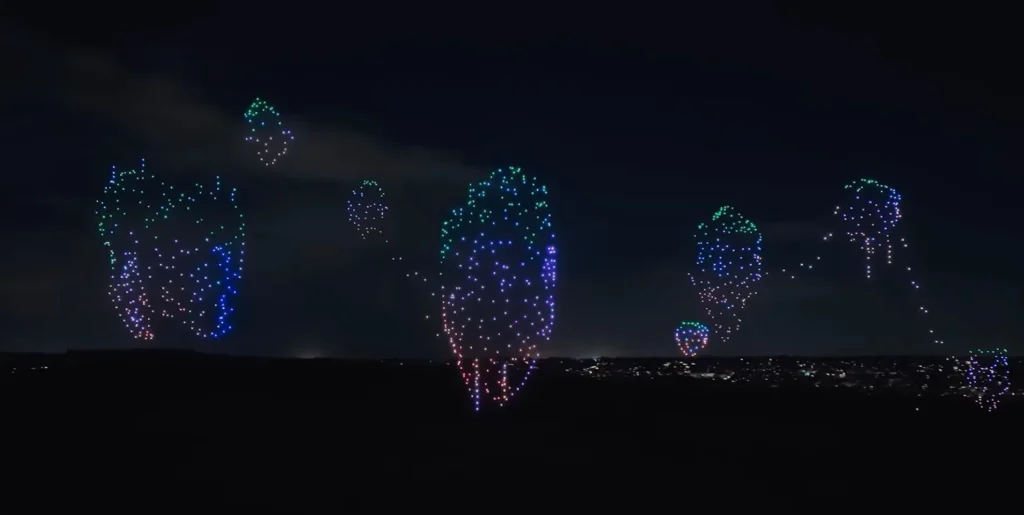 Une scène nocturne montre plusieurs grandes formations de drones multicolores dans le ciel, disposés en forme de ballon au-dessus du Parc Walt Disney Studios pendant le spectacle de drones Avatar, avec les lumières de la ville au loin qui scintillent en dessous.