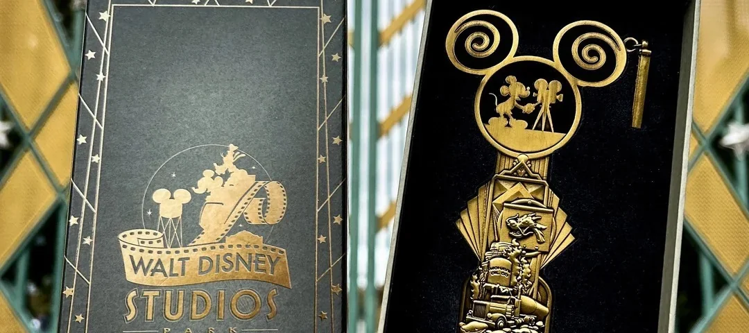 Une main tient une boîte Walt Disney Studios Édition Limitée et un grand Clé Collector Jumbo devant la porte d'entrée emblématique du studio.