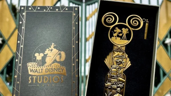 Une main tient une boîte Walt Disney Studios Édition Limitée et un grand Clé Collector Jumbo devant la porte d'entrée emblématique du studio.