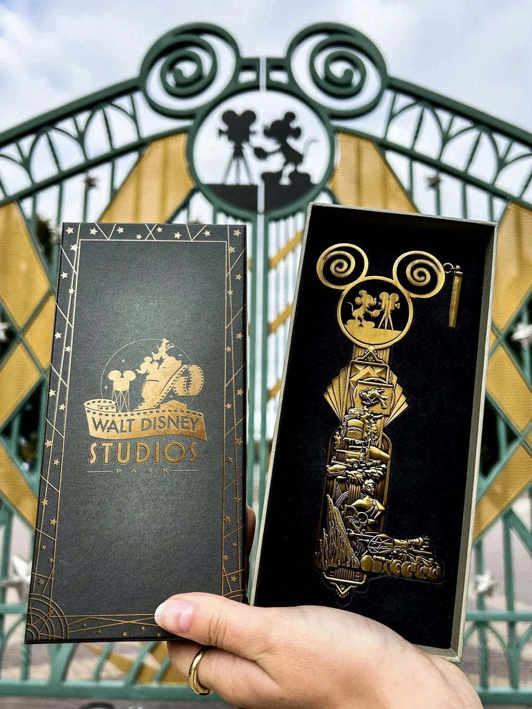 Une main tient une boîte Walt Disney Studios Édition Limitée et un grand Clé Collector Jumbo devant la porte d'entrée emblématique du studio.