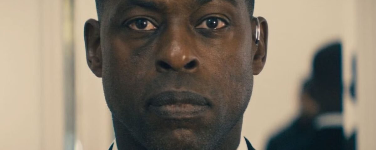 Un homme en costume à l'expression sérieuse se tient à l'intérieur, regardant directement la caméra, comme s'il faisait partie d'une bande-annonce captivante de Disney+ Paradise Saison 2.