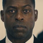 Un homme en costume à l'expression sérieuse se tient à l'intérieur, regardant directement la caméra, comme s'il faisait partie d'une bande-annonce captivante de Disney+ Paradise Saison 2.