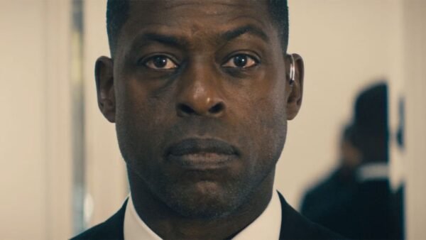 Un homme en costume à l'expression sérieuse se tient à l'intérieur, regardant directement la caméra, comme s'il faisait partie d'une bande-annonce captivante de Disney+ Paradise Saison 2.