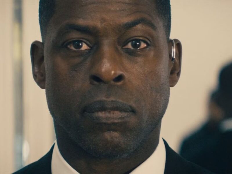 Un homme en costume à l'expression sérieuse se tient à l'intérieur, regardant directement la caméra, comme s'il faisait partie d'une bande-annonce captivante de Disney+ Paradise Saison 2.