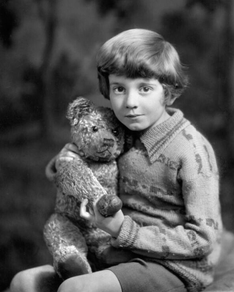 Un jeune enfant aux cheveux courts, assis, tenant un ours en peluche usé rappelant Winnie l'Ourson, vêtu d'un pull à motifs et d'un short, pose devant un fond de studio flou.